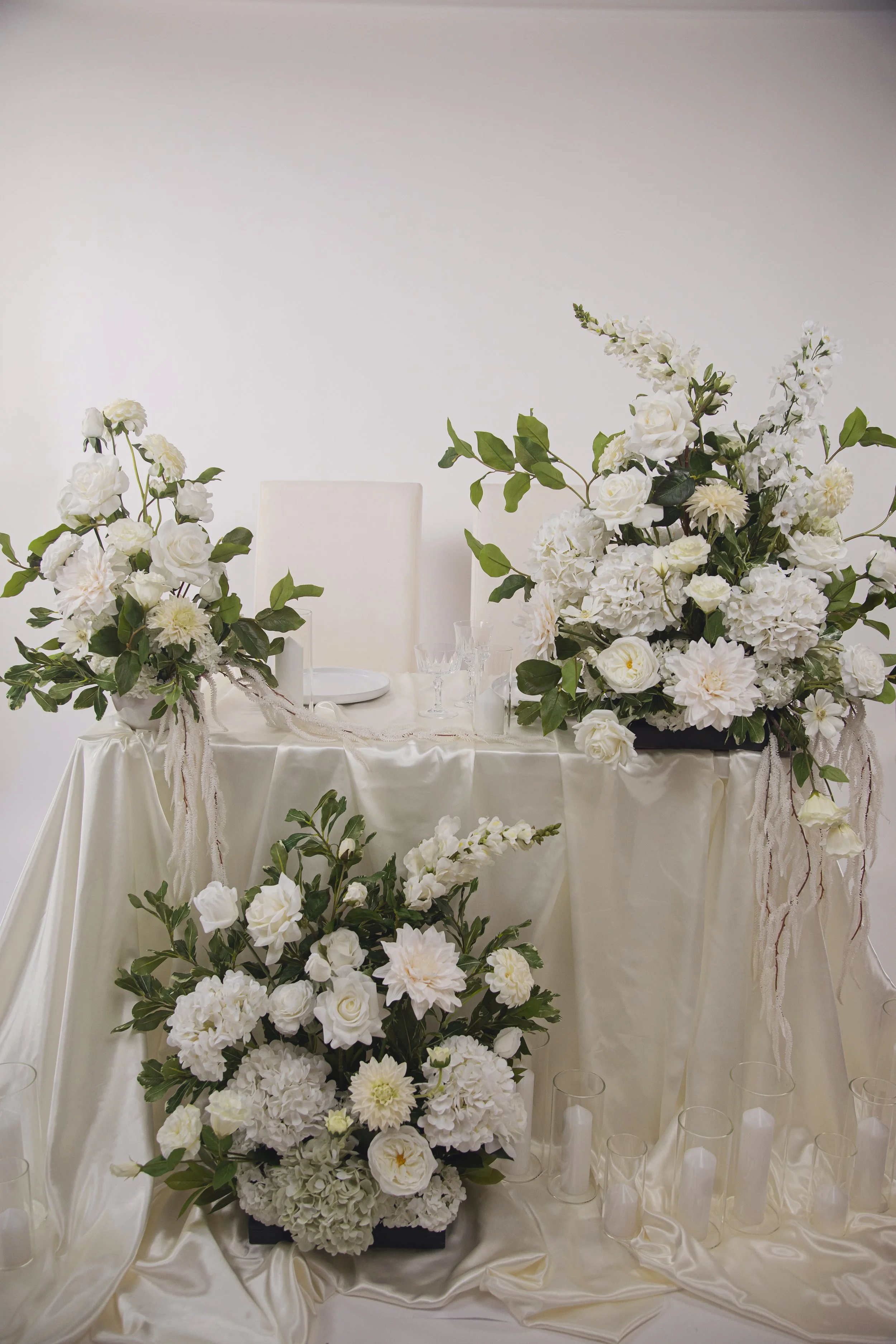 Elegant wedding table decorated with large arrangements of white roses, hydrangeas, and other white flowers, with candle holders on a satin tablecloth.