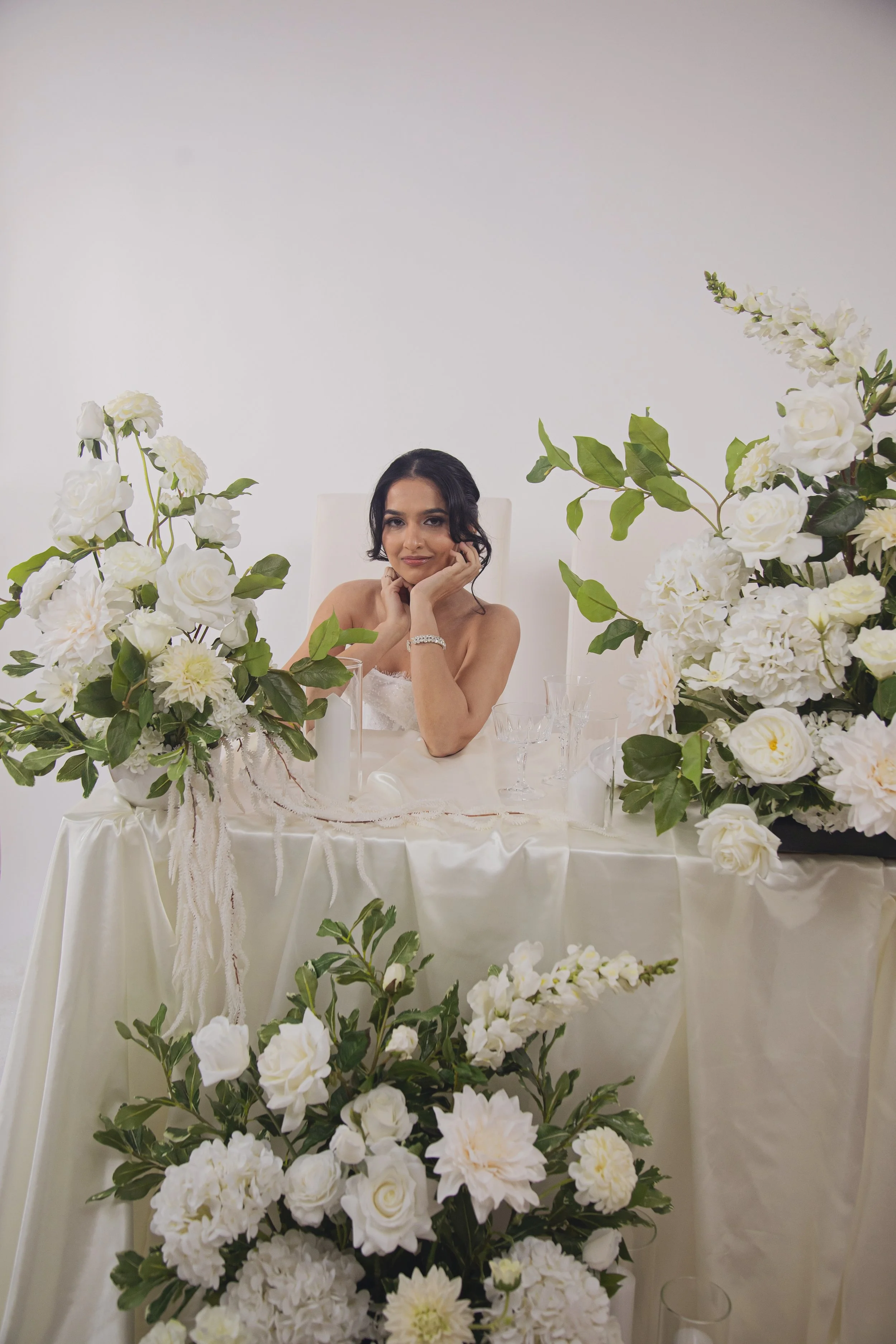 A woman with dark hair in an elegant updo sits at a beautifully decorated table with white flowers and greenery, wearing a strapless white dress, and resting her chin on her hand.