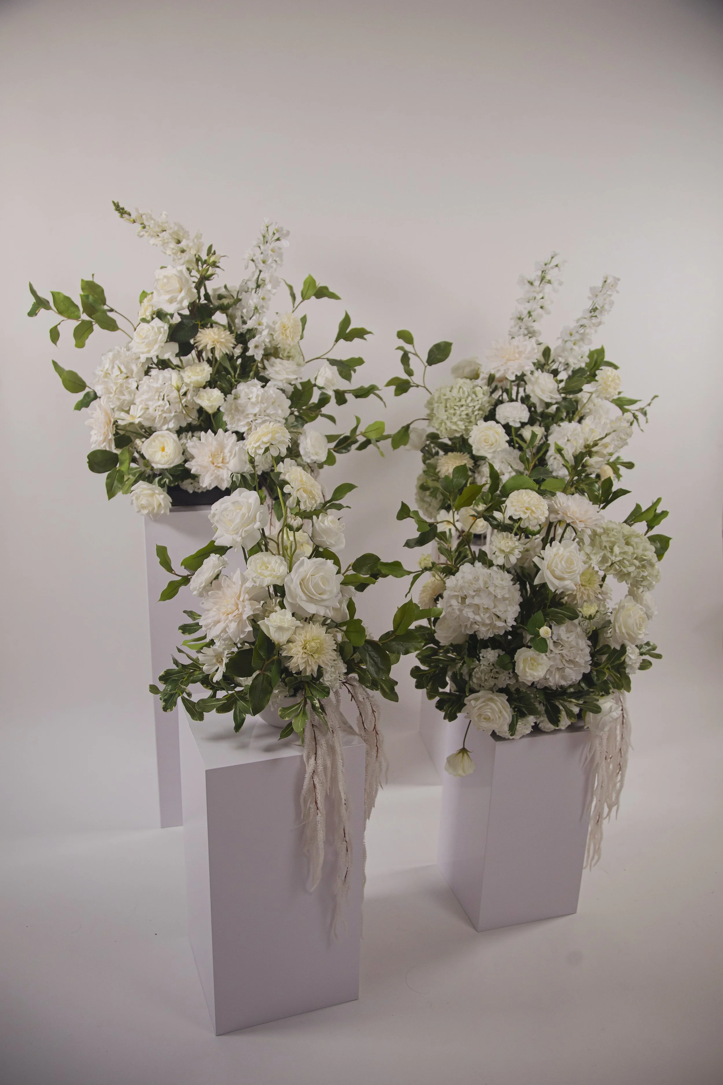 Three white flower arrangements in white rectangular vases on white pedestals against a white background.