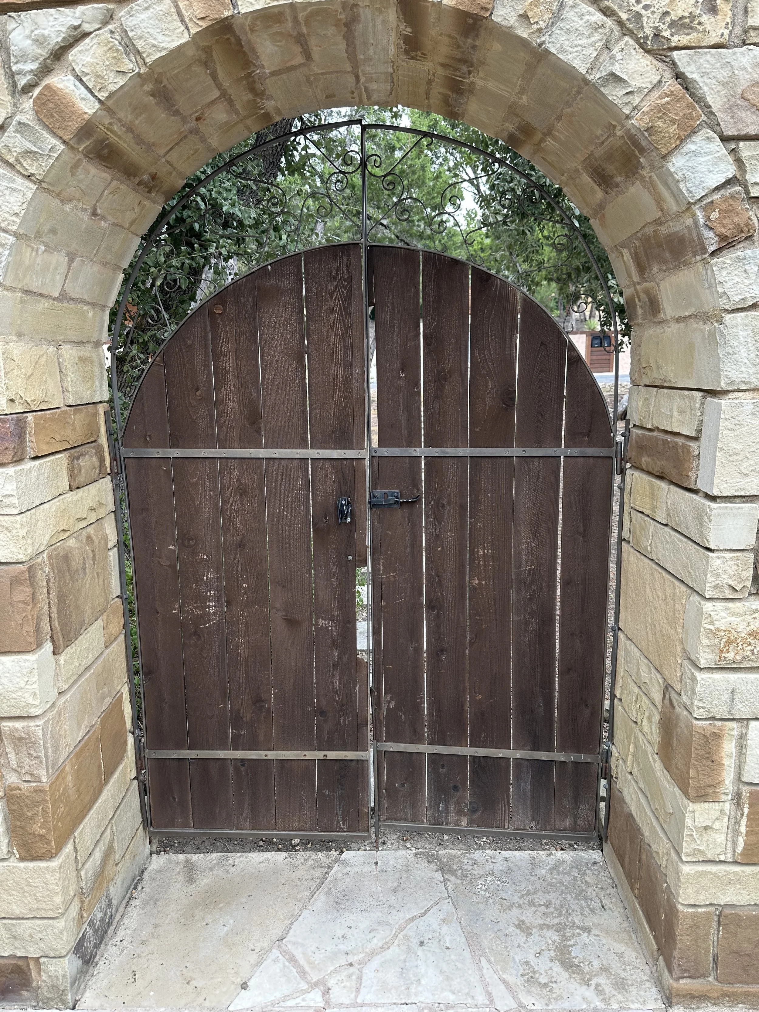 Wooden gate with metal hardware set in a stone archway, with trees and greenery visible through the gate.