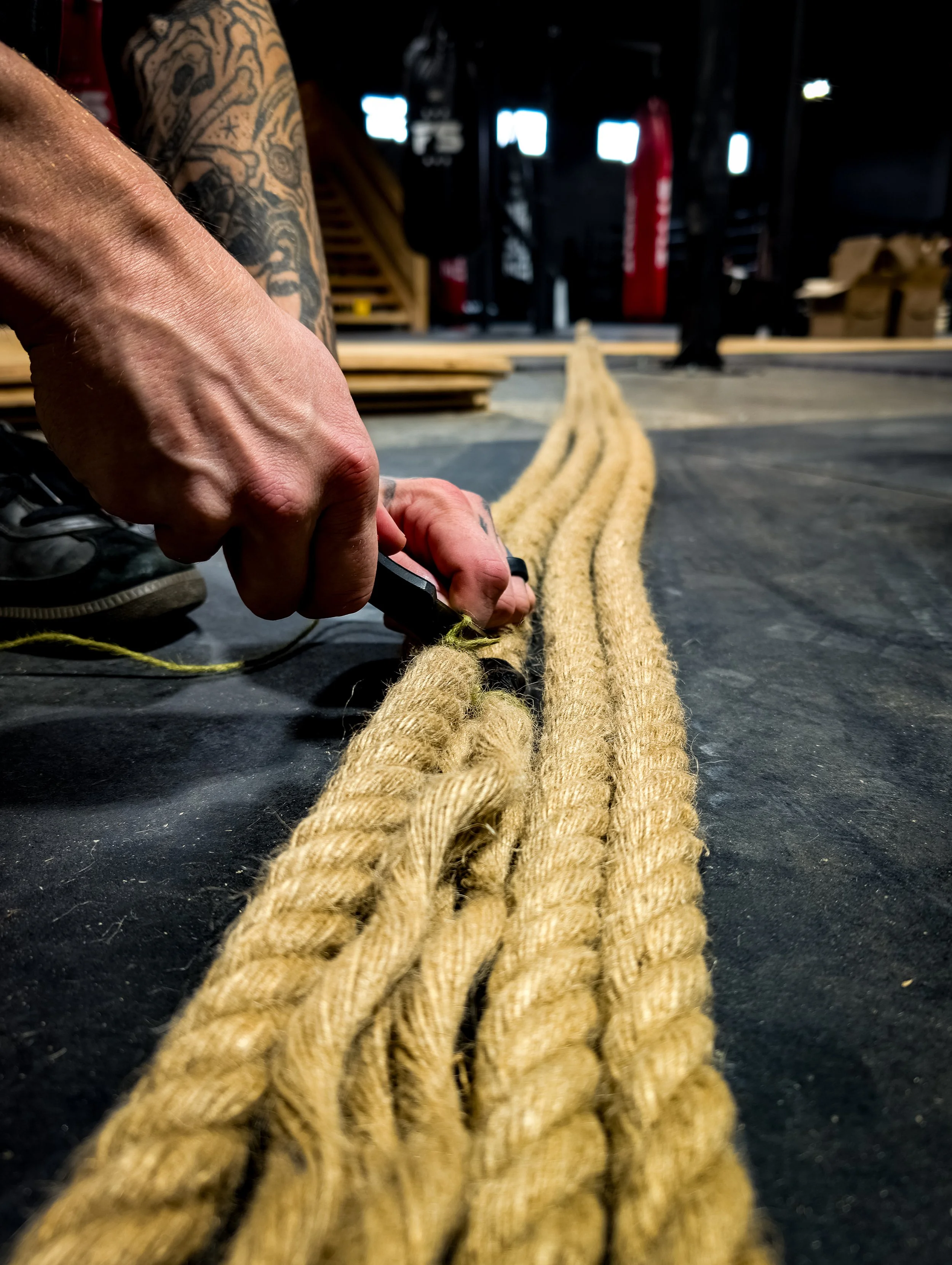 A person with tattoos is cutting ropes on a gym floor before installing on a custom boxing ring.