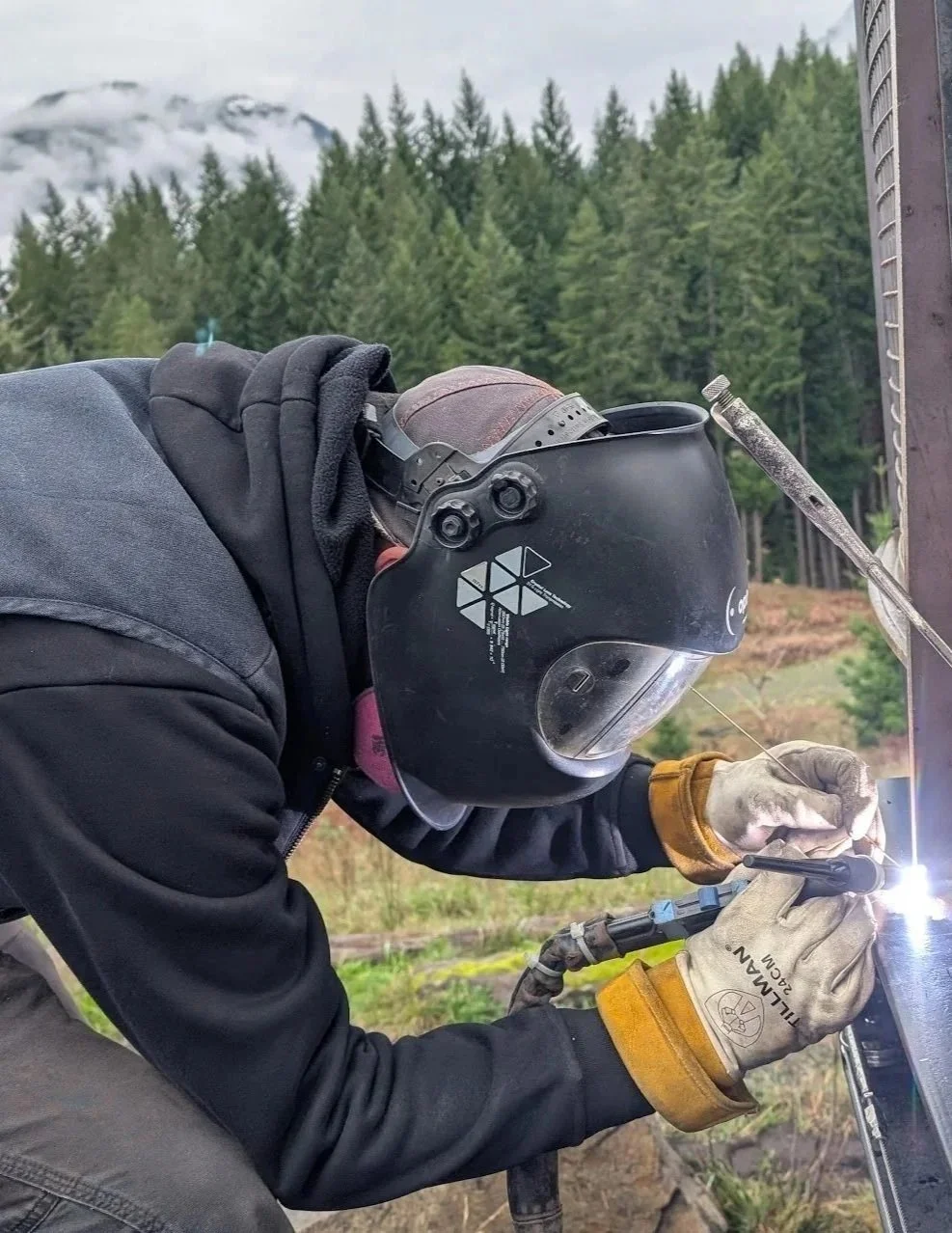 A person welding outdoors in a forested area, wearing a welding helmet, gloves, and protective clothing, using a welding torch to work on metal.