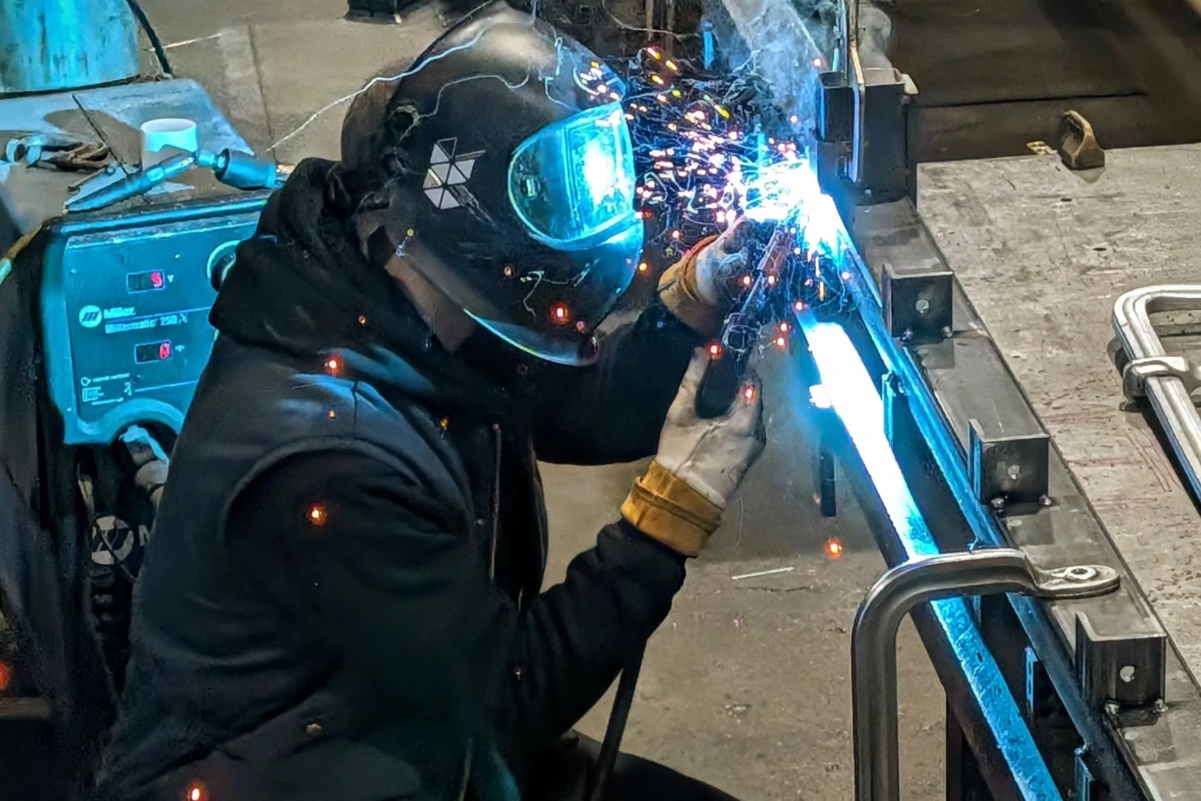 A person welding metal at a workbench, wearing a protective helmet, gloves, and a black jacket. Sparks and bright light from the welding process are visible.