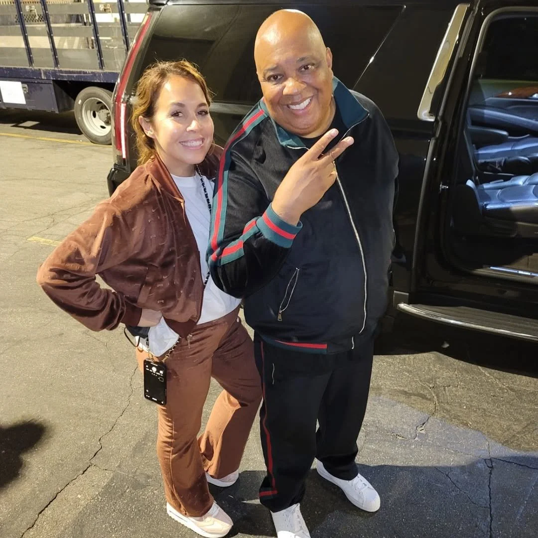 A woman and a man posing for a photo in a parking lot at night. The woman is wearing a brown jacket, brown pants, and white sneakers with a cell phone clipped to her pocket. The man is dressed in a black tracksuit with red and green accents, white sn