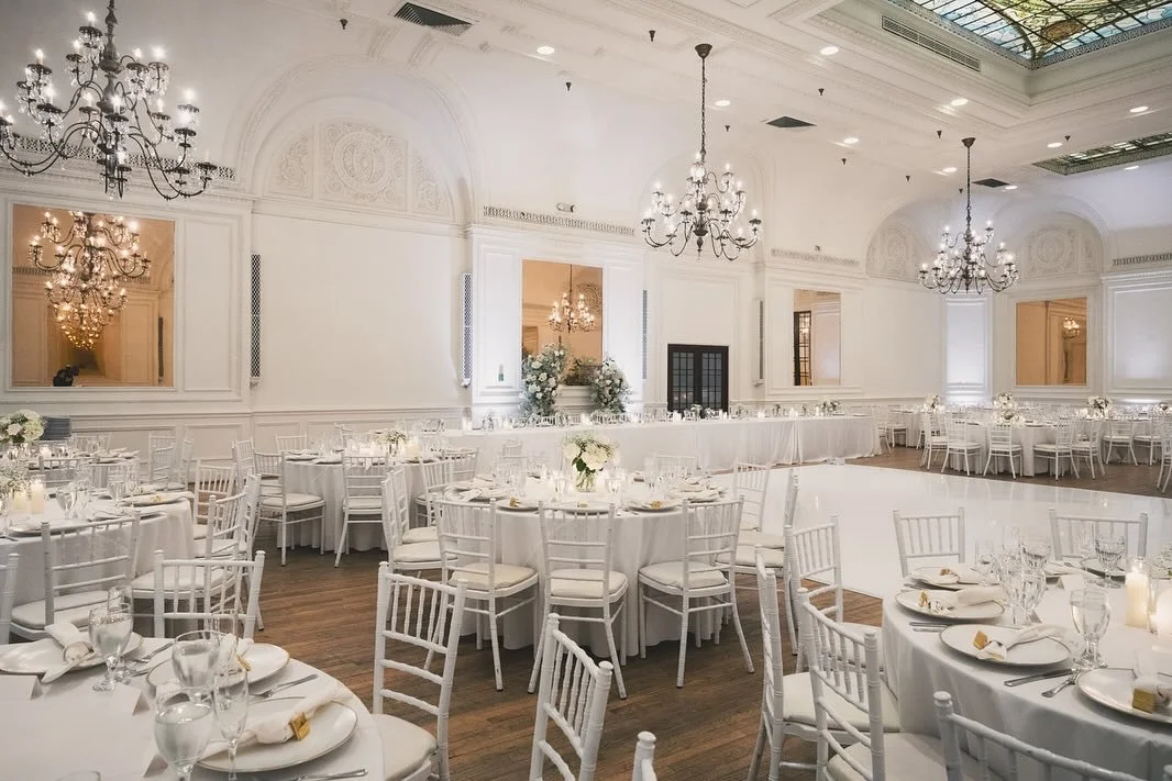 Elegant banquet hall decorated for a wedding or formal event with white drapery, chandeliers, and round tables set with white tablecloths, flowers, and glassware.