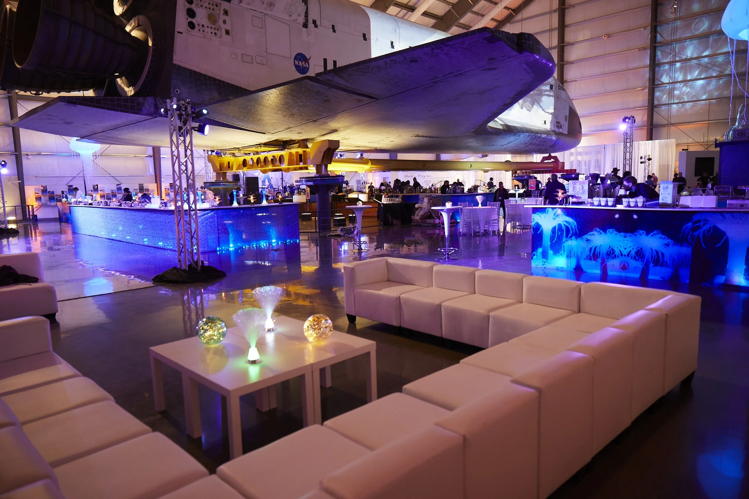 Interior of a space-themed lounge or event space with a space shuttle hanging from the ceiling, illuminated bar areas with blue lighting, and seating arrangements including white couches and tables with decorative lighting.