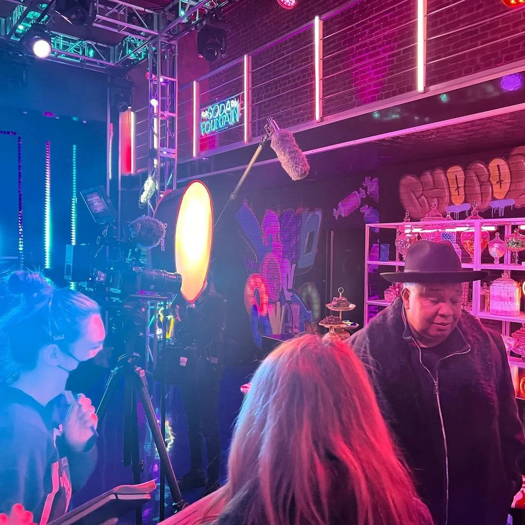 People in a bar with colorful neon lights, including a neon sign that reads 'Soda Fountain', and a camera crew filming the scene.