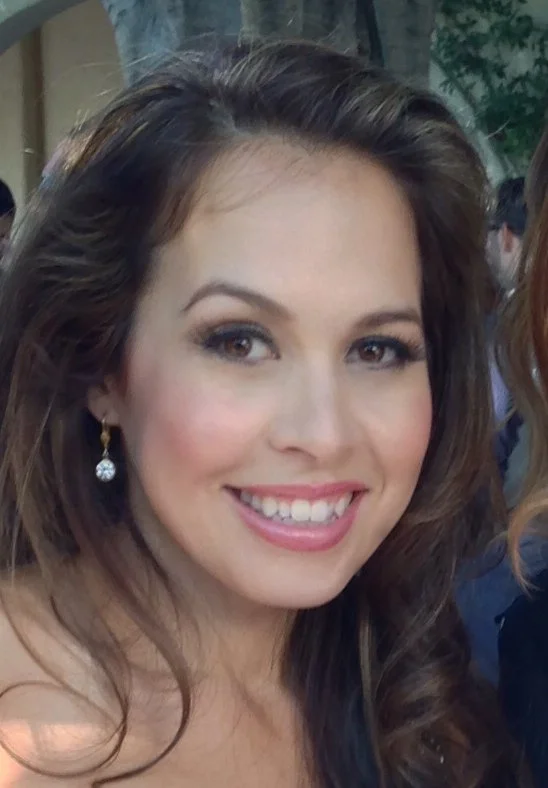 Close-up of a smiling woman with wavy brown hair, wearing pearl earrings, in an outdoor setting.