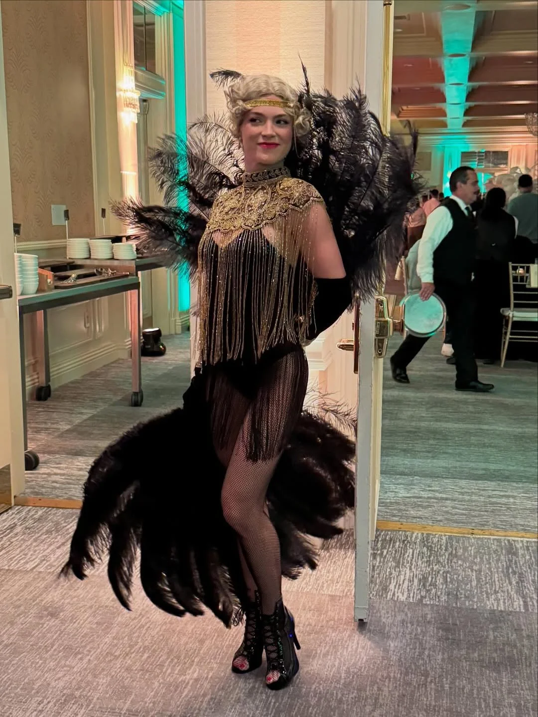 A woman dressed in a flapper-style costume with black feathers, gold lace, and fringe, wearing fishnet stockings and black lace-up high heels, standing indoors at a lively event.