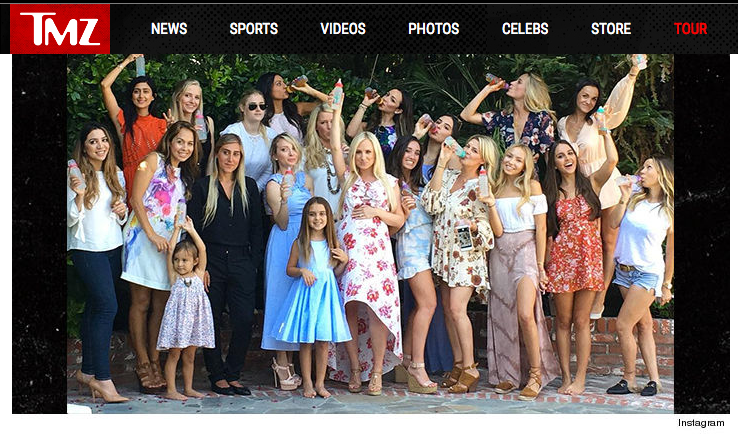 A group of women and girls gathered outdoors for a celebration, some holding drinks and posing for a photo, with lush greenery in the background.