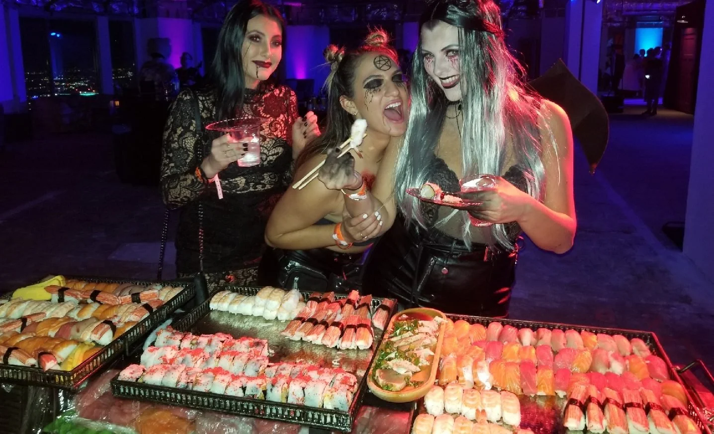Three women in a dimly lit setting dressed in Halloween costumes, smiling and enjoying sushi with drinks on a table.