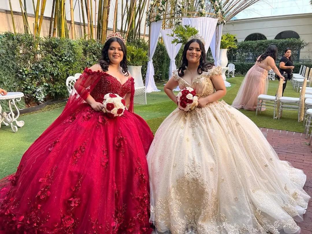 Two women in formal gowns holding bouquets, standing outdoors on a lawn with a decorated arch in the background. One woman is in a red gown with floral embroidery, the other in a cream-colored gown with lace details.