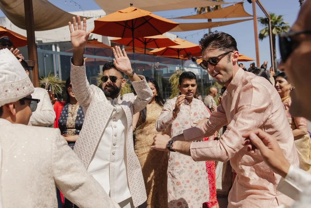 People dancing and celebrating outdoors at a daytime event, possibly a wedding or cultural festival, with orange umbrellas and palm trees in the background.