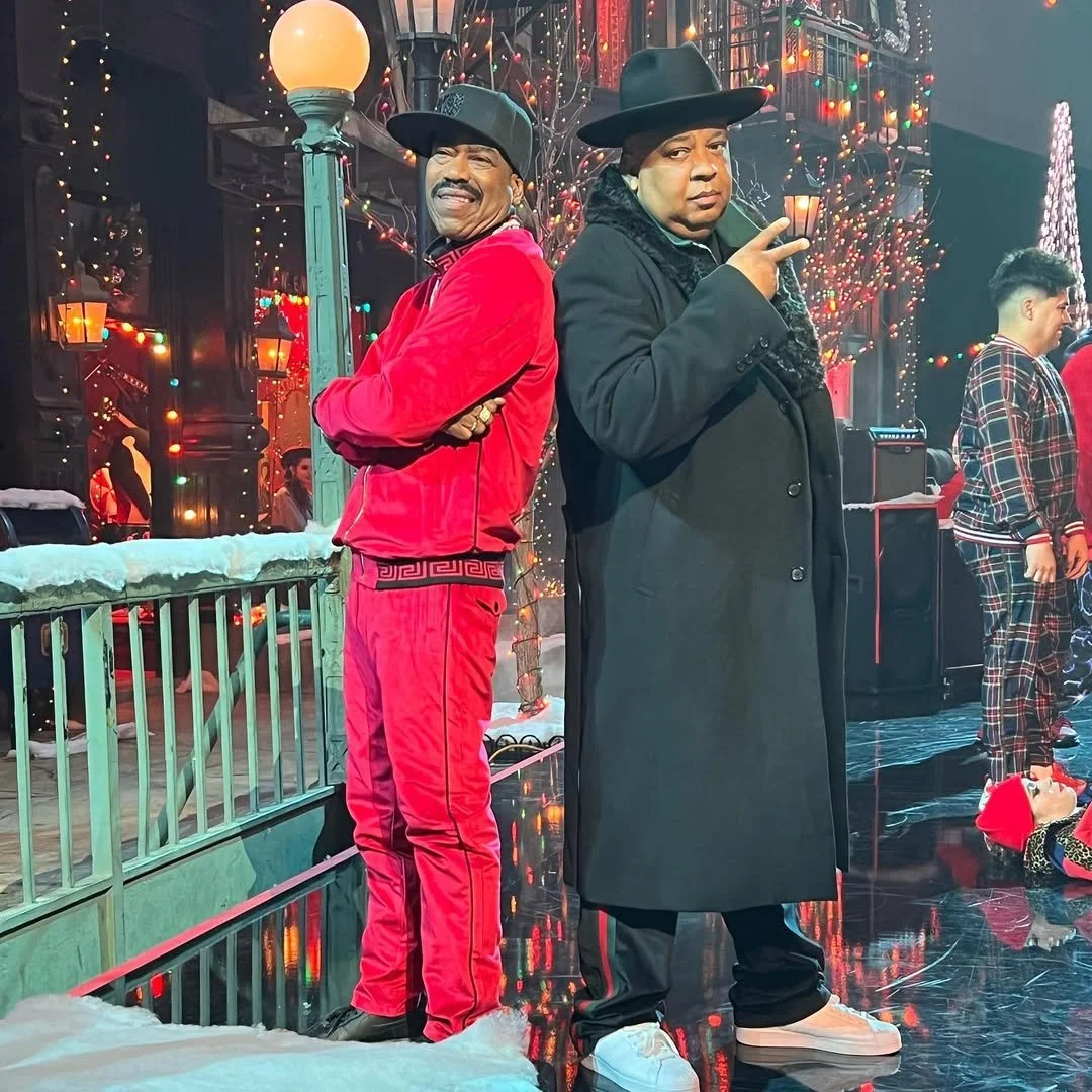 Two men dressed in costumes posed back-to-back in a festive setting with Christmas lights. One man wears a red velvet tracksuit with black trim, and the other wears a black coat and wide-brimmed black hat, making a gesture with his hand.