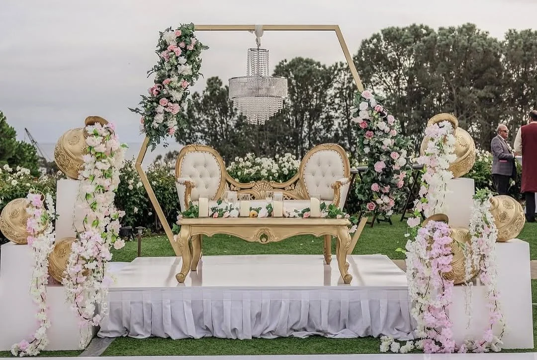 Wedding stage with gold and white furniture, floral decorations, and a chandelier in an outdoor setting.