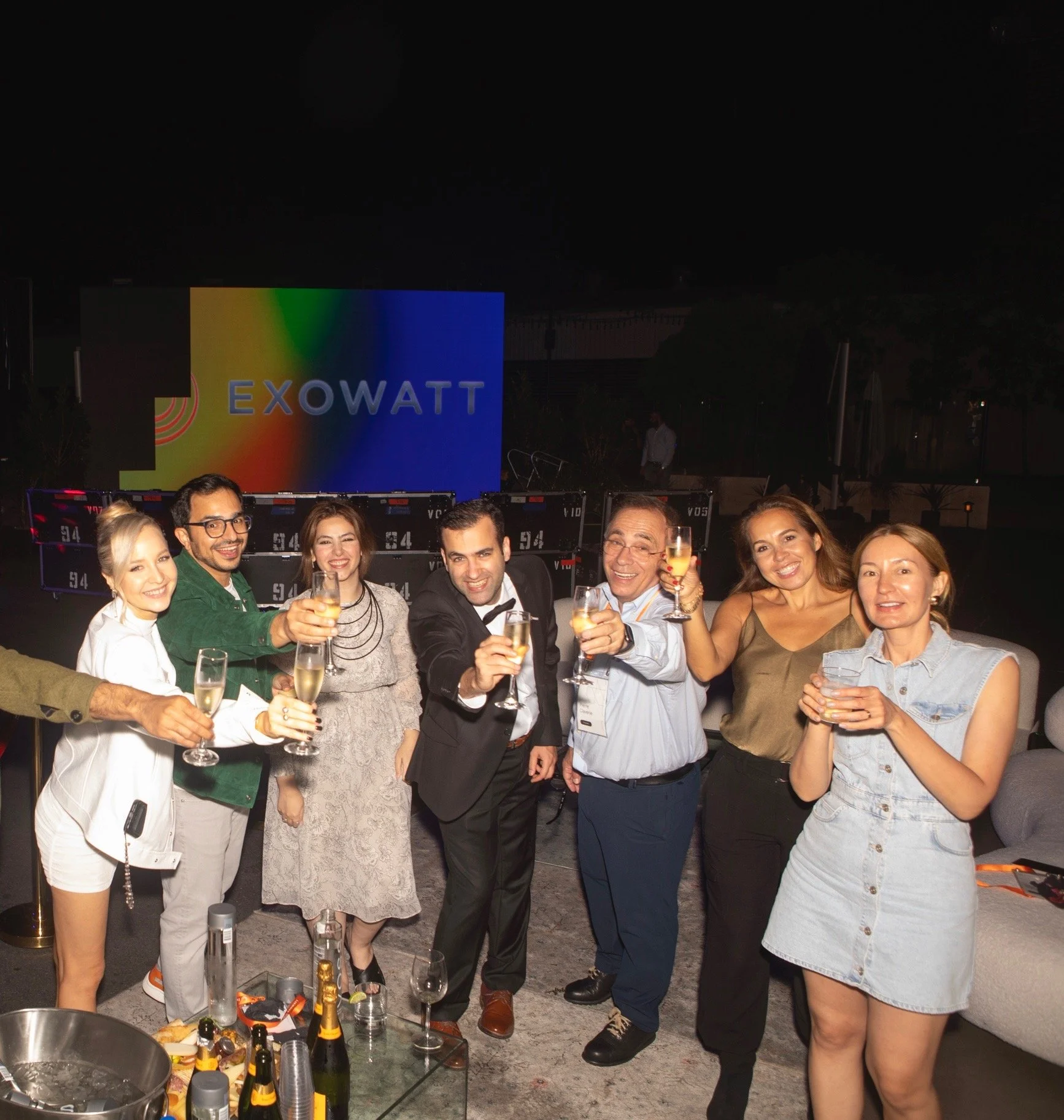 Group of seven people celebrating with champagne glasses at a party with a sign that says EXOWATT in background.