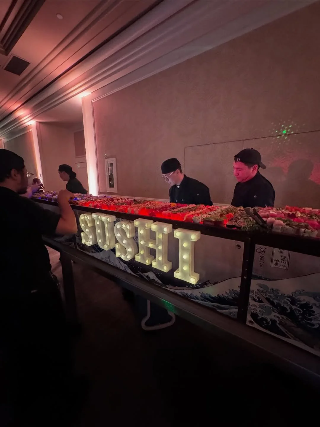 A sushi bar with a lit sign spelling 'SUSHI' in front of a buffet of assorted sushi, with three staff members behind the counter preparing food, and customers selecting sushi in a dimly lit room.