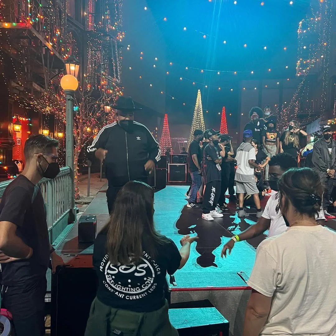 People dancing and socializing on a decorated stage with Christmas trees and string lights, some wearing masks, in a festive indoor setting.