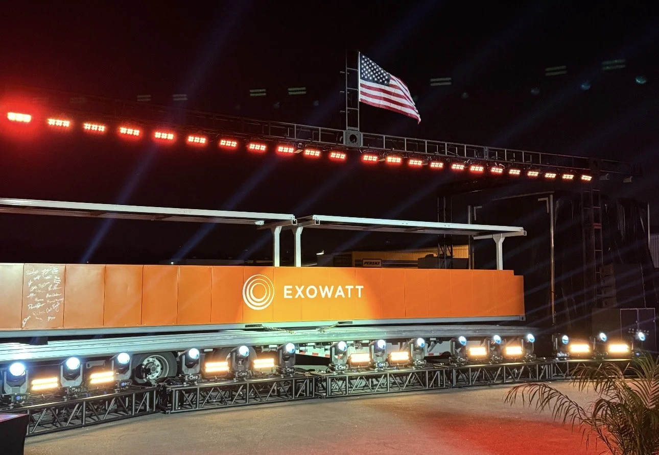 Stage setup with orange panel displaying 'EXOWATT' and an American flag flying above, illuminated by stage lights and red overhead lighting.