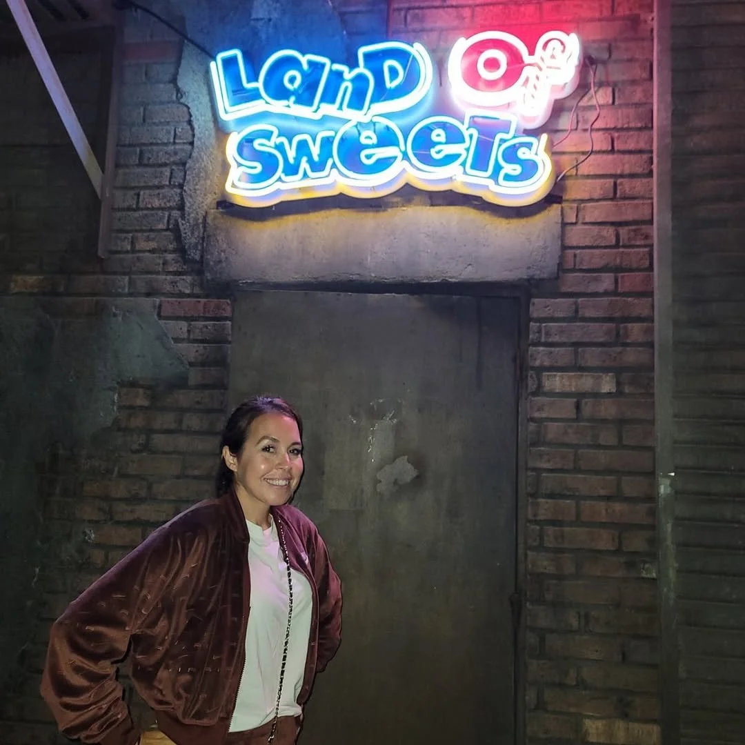 A smiling woman in a white shirt and brown jacket standing in front of a brick wall at night, with a neon sign above her that says 'Lana Sweets' and features a small butterfly design.