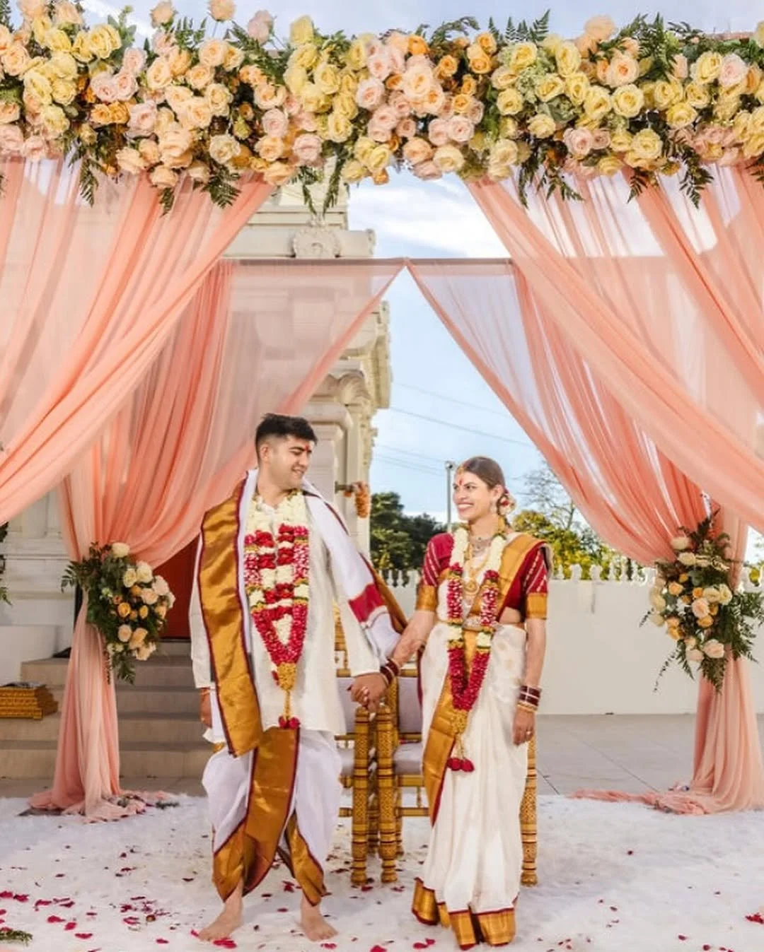 A couple in traditional Indian wedding attire holding hands under a floral canopy with pink drapes, outdoors.