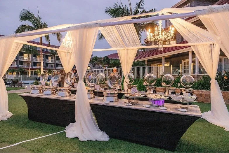 Buffet table set up under a white canopy with draped curtains, featuring chafing dishes and a purple lit item, on a lawn with palm trees and a building in the background during evening.