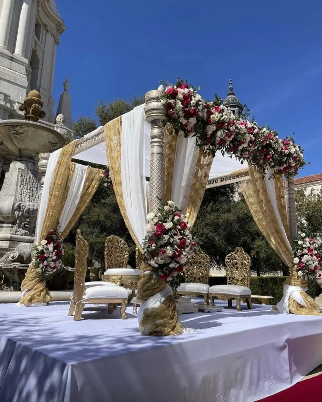 A decorated wedding stage with white and gold drapes, floral arrangements of pink, white, and red roses, and ornate chairs, set outdoors under clear blue sky and historic architecture.