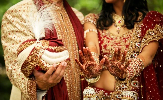 Indian bride and groom dressed in traditional wedding attire, showing henna-decorated hands and the groom holding a feathered ceremonial item.