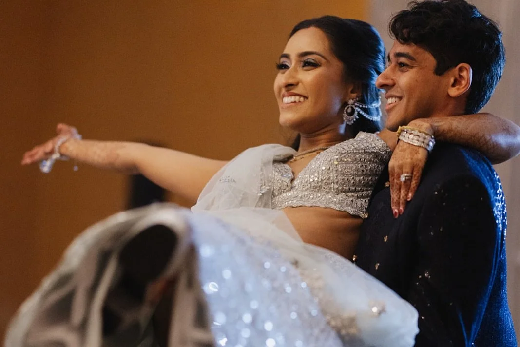 A joyful couple dancing at a wedding celebration, the woman wearing traditional Indian attire and jewelry, and the man in a dark suit, both smiling and looking happy.