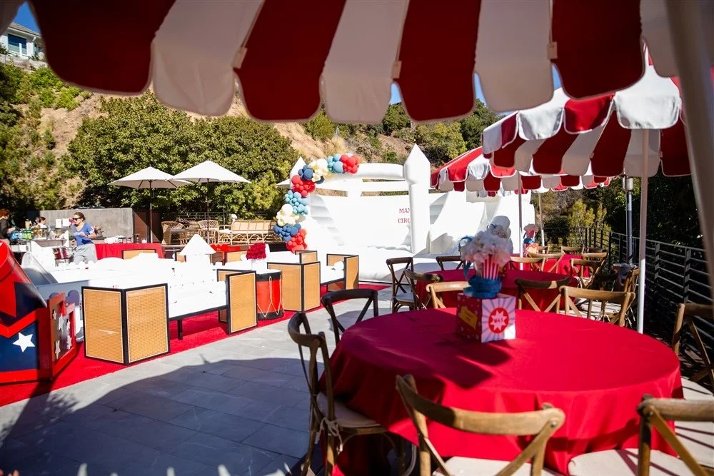 Outdoor area decorated for a circus-themed event with red and white striped umbrellas, round tables with red tablecloths, chairs, balloon decorations, and an inflatable circus tent in the background.