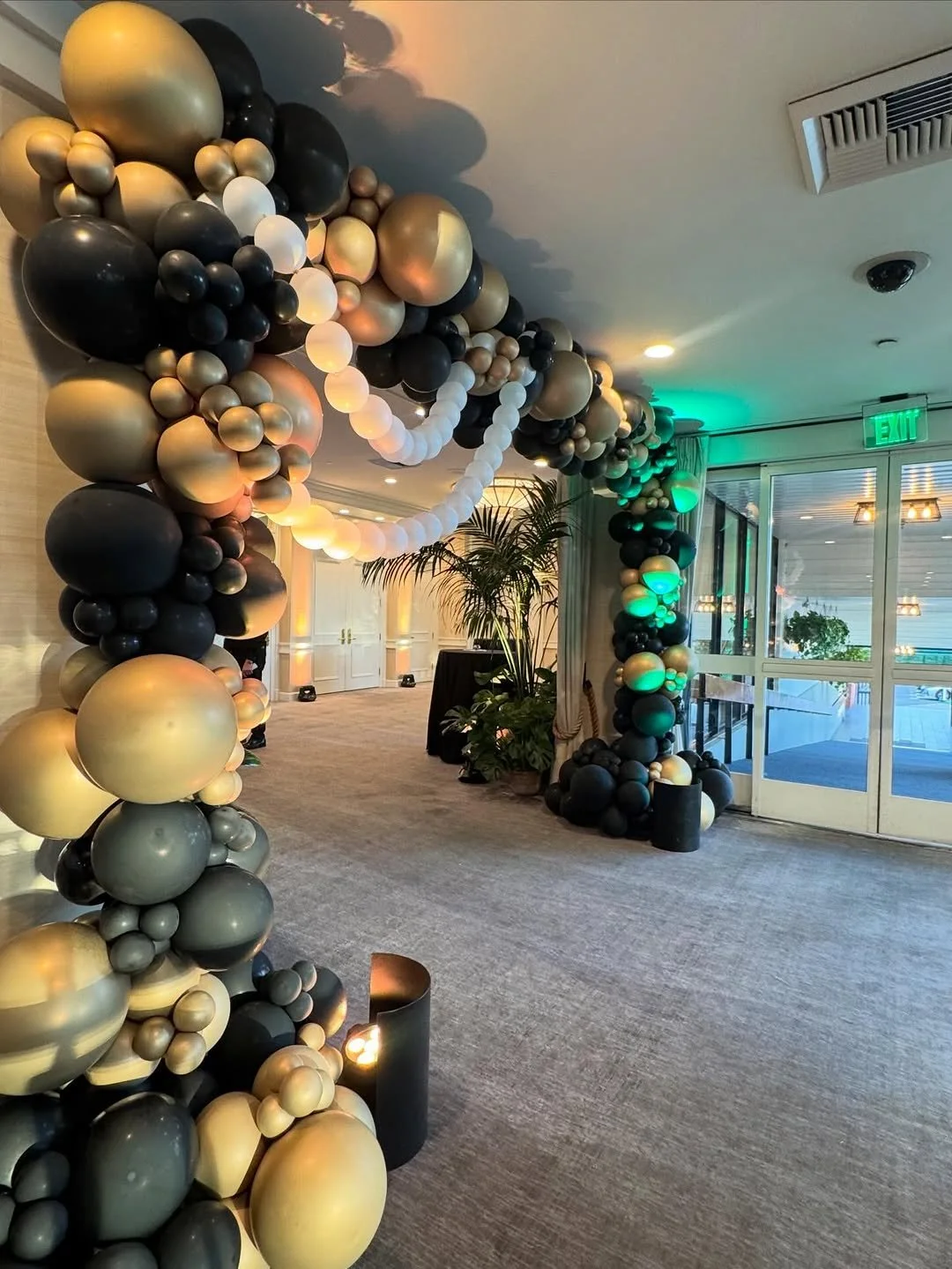 Decorative arch made of black, white, gold, and beige balloons at an indoor event venue near glass double doors and potted plants.