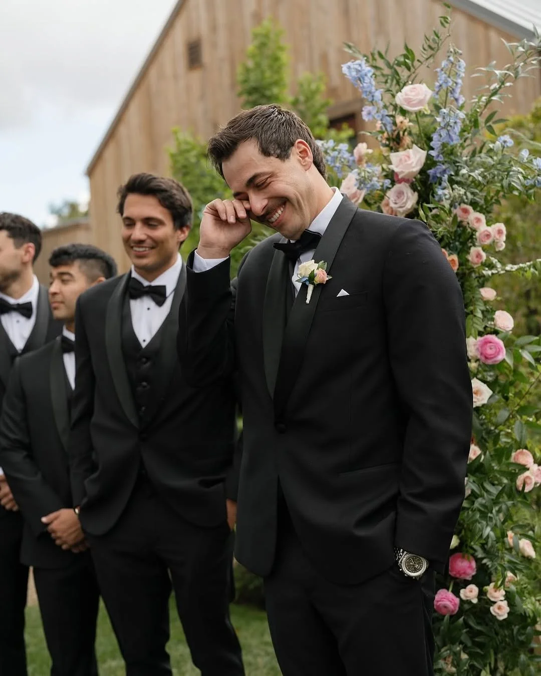A group of men dressed in black tuxedos with bow ties, standing outdoors near floral arrangements and wooden building, with one man smiling and touching his face, possibly during a wedding celebration.