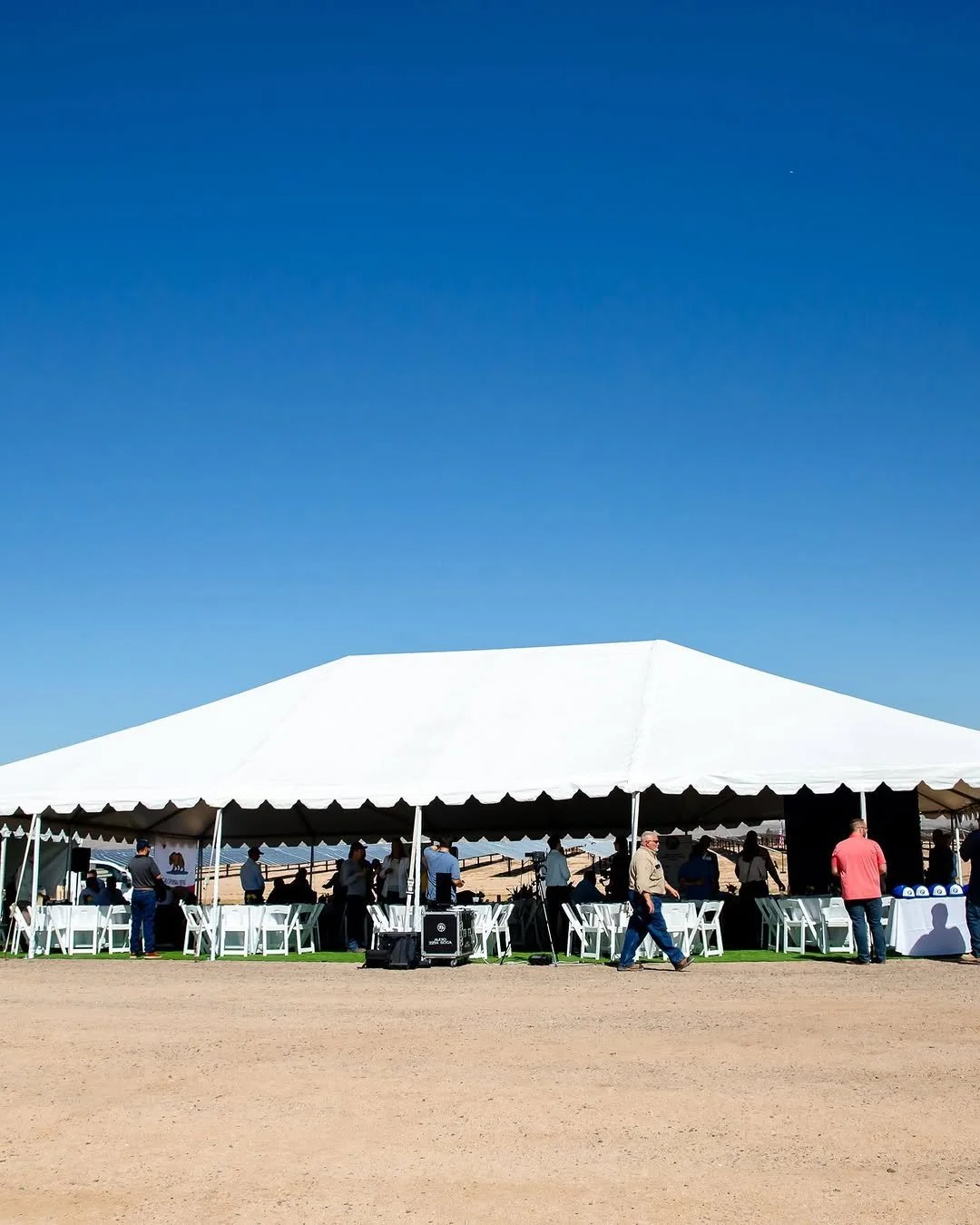 A large white event tent set up outdoors on a clear day with people standing and sitting around. The tent is on a dirt or sandy surface under a bright blue sky.