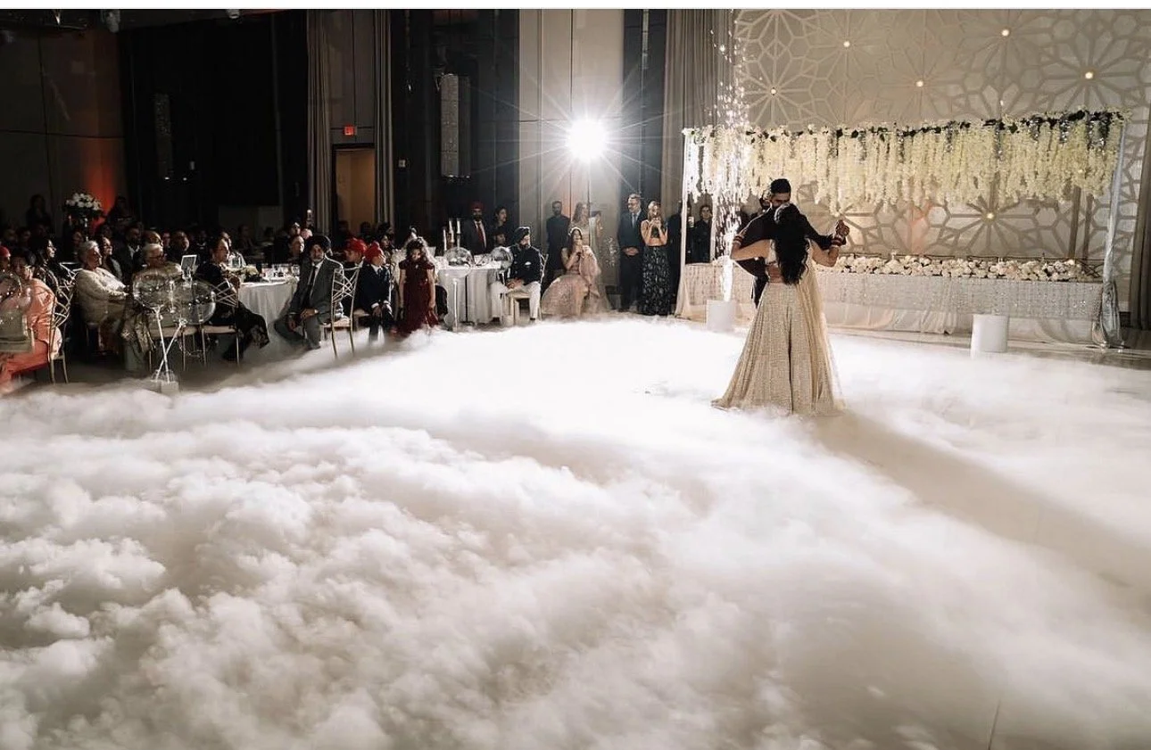 Couple dancing at their wedding reception with guests seated at tables and standing around, decorated with white flowers. Fog covers the dance floor, and a bright light illuminates the scene.