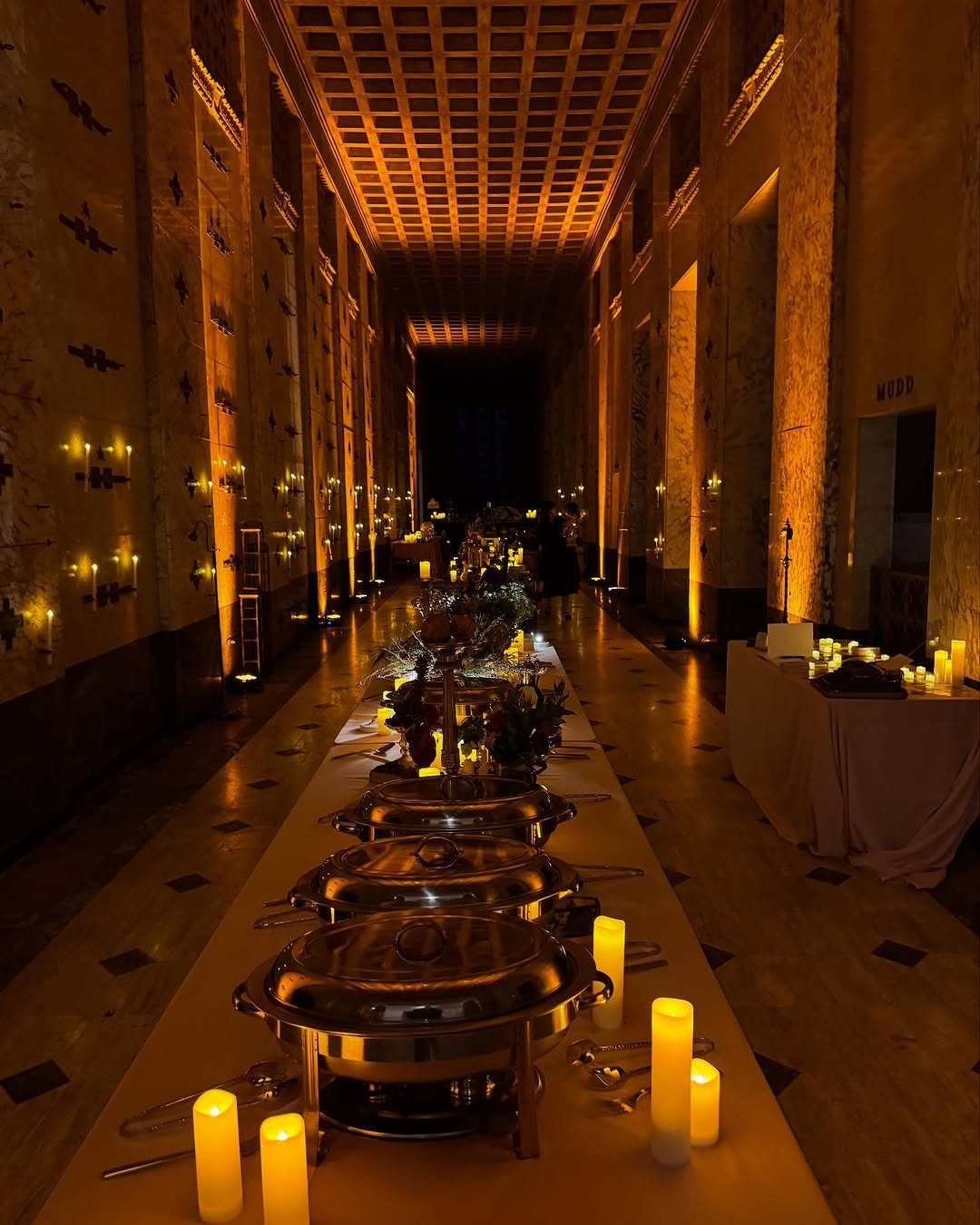 A dimly lit banquet hall with a long table set for an event, illuminated by candles and decorative lights on the walls.