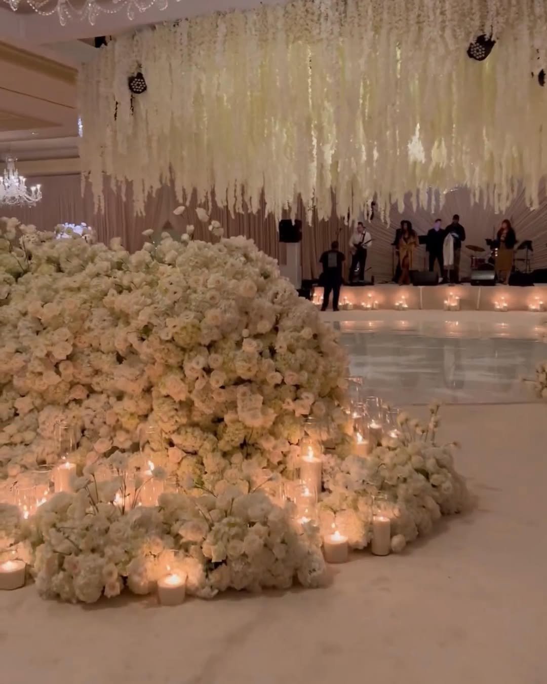 Elegant wedding reception with a large floral centerpiece made of white flowers, surrounded by candles and soft lighting, with a band performing on stage in the background.
