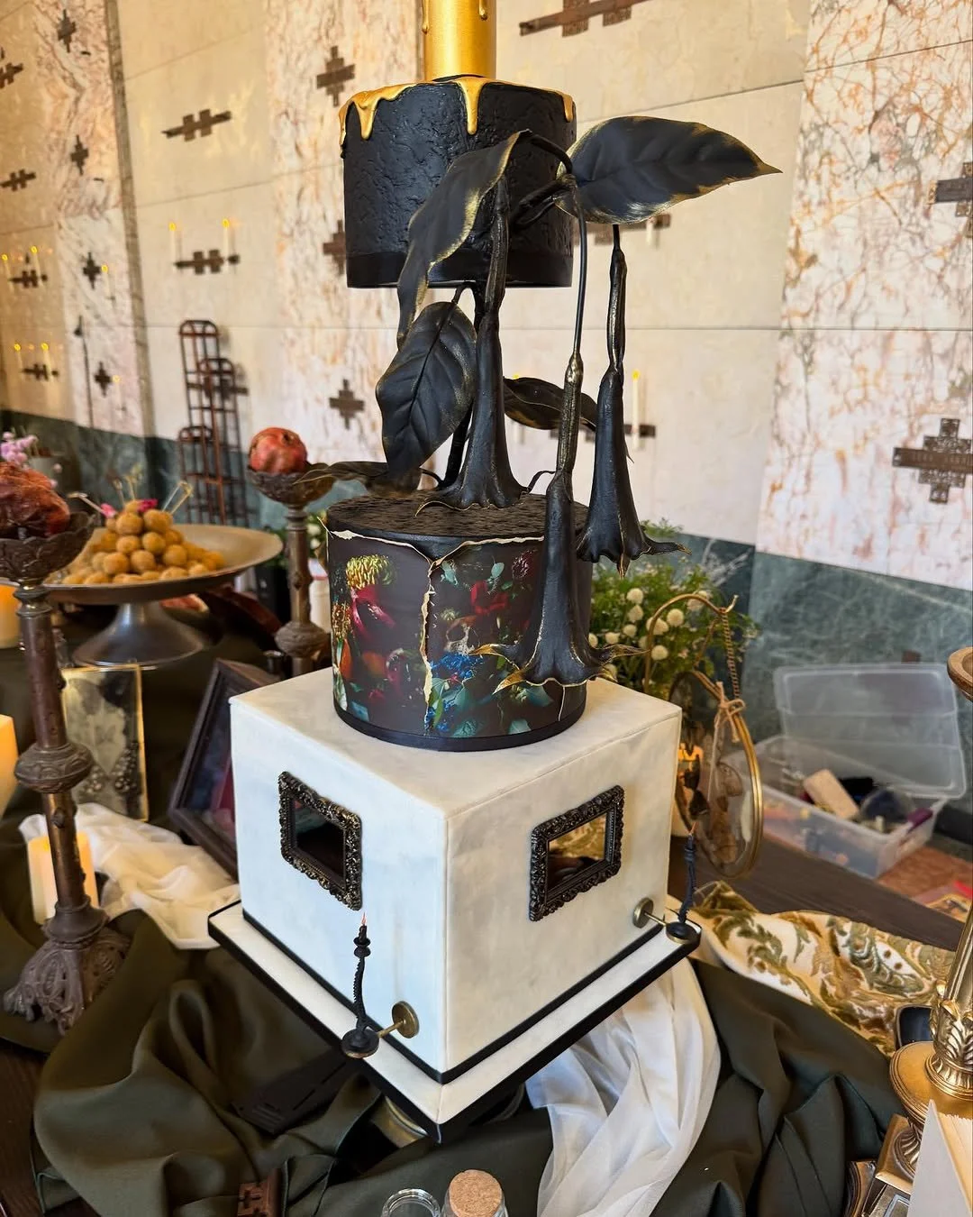 A decorative sculpture of a black cake with hanging black leaves and a gold top, placed on a white pedestal among various vintage and ornate decorative items.