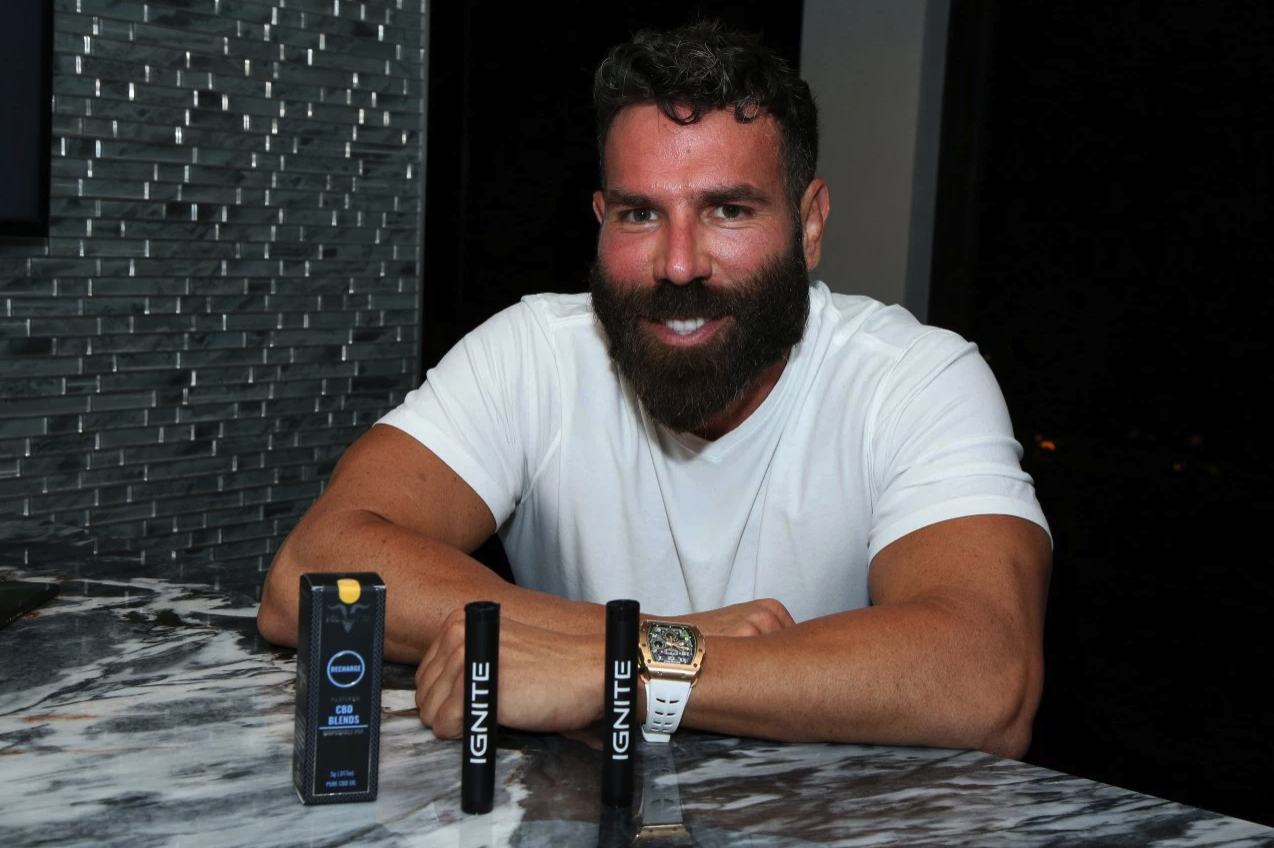 A bearded man in a white t-shirt sitting at a marble table, smiling, with two Ignite CBD vape products and a box on the table in front of him.