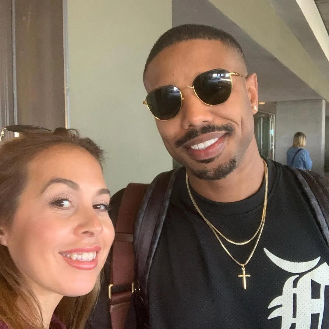 A woman with light skin and brown hair, and a man with dark skin wearing sunglasses, standing close together and smiling for a photo indoors.