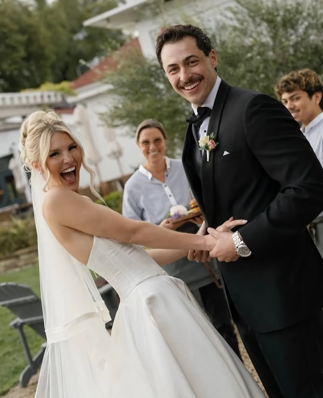 A bride and groom are holding hands and smiling at their wedding outdoors, with three guests in the background.
