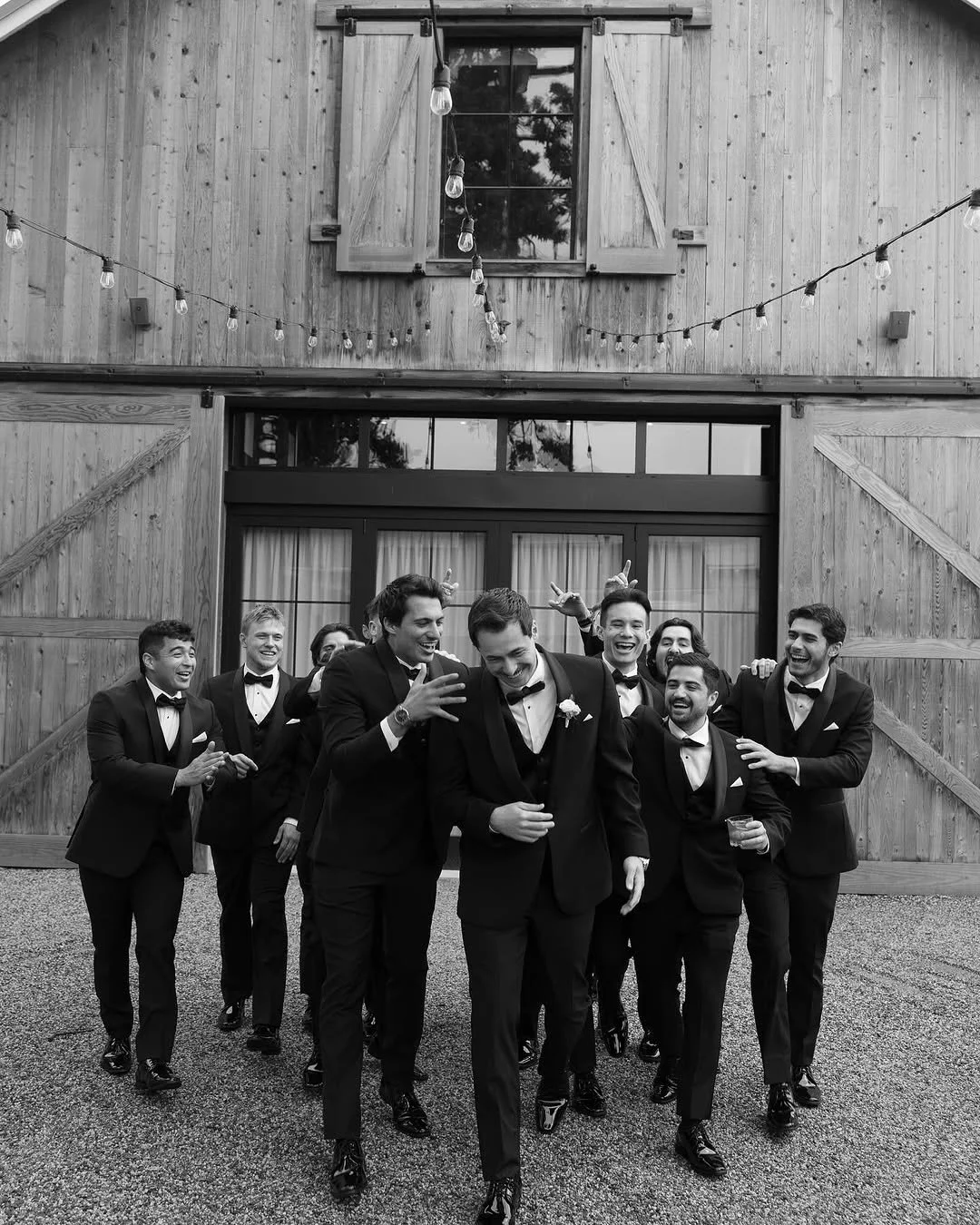 A group of men dressed in tuxedos smiling and laughing, walking outside a wooden building with string lights overhead. One man in front is holding a glass. They appear to be celebrating.