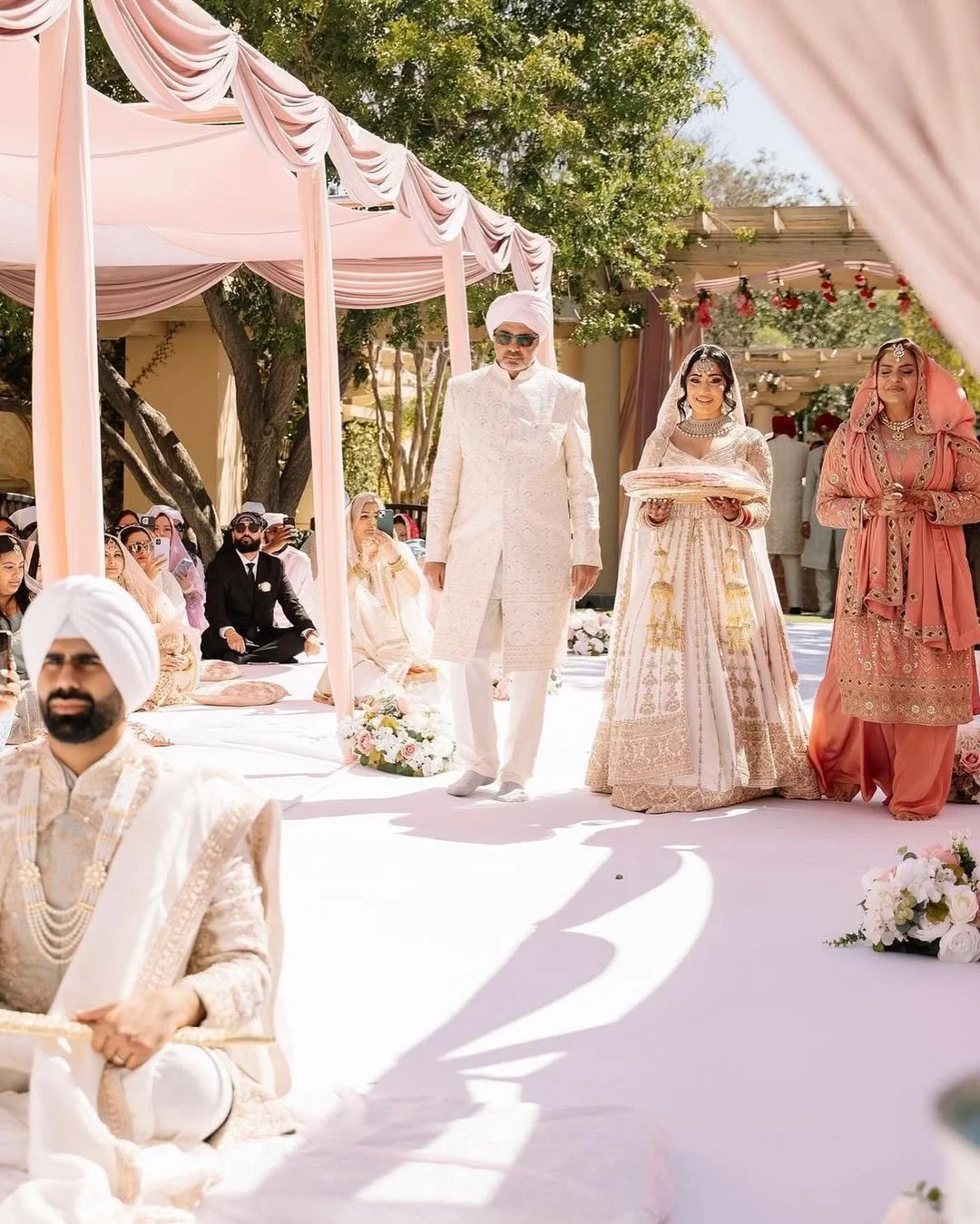 Indian wedding ceremony with a groom in white traditional attire and turban, two women in sarees or lehengas, and guests seated on the ground under pink draped canopy and floral decorations.