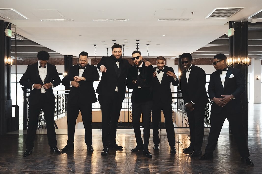 Group of seven men in tuxedos with bow ties, posing together indoors against railing, with wooden floors and large windows.