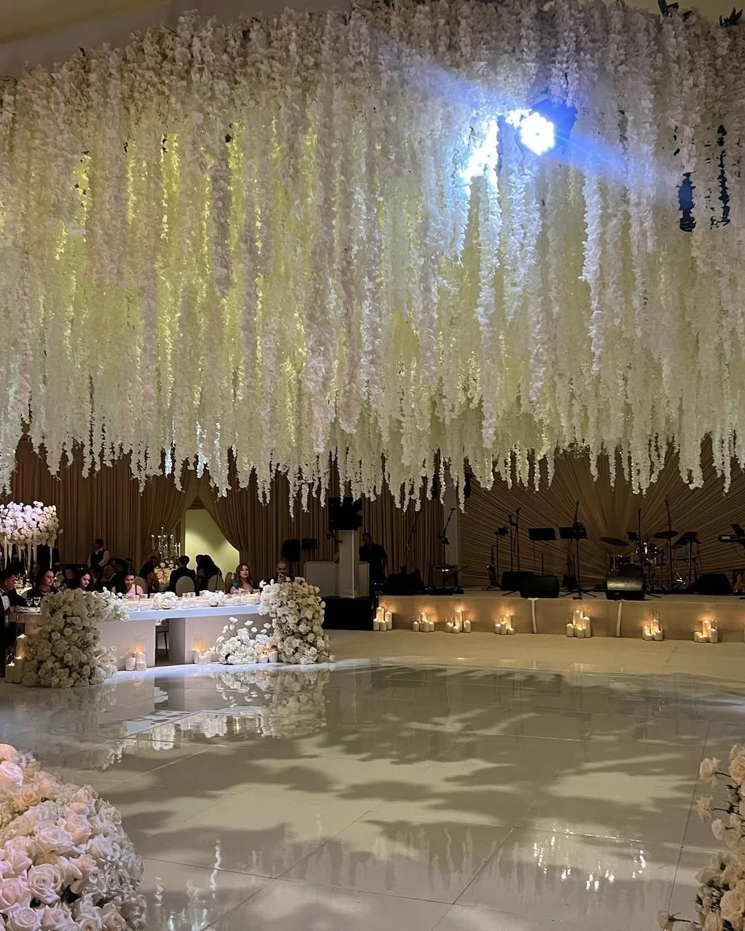 Wedding reception with hanging white flower decorations, a long table with floral arrangements, and candles on the floor. Soft lighting and a stage with musical instruments in the background.