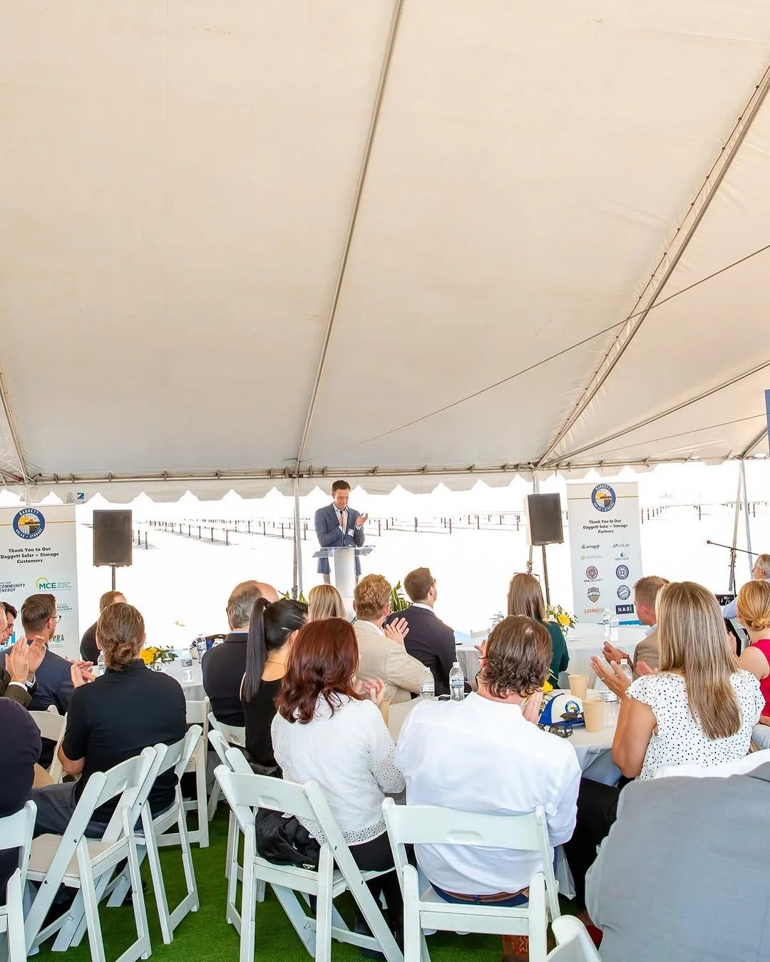 Business conference under a large white tent with a speaker at a podium and an audience seated at round tables, some clapping.