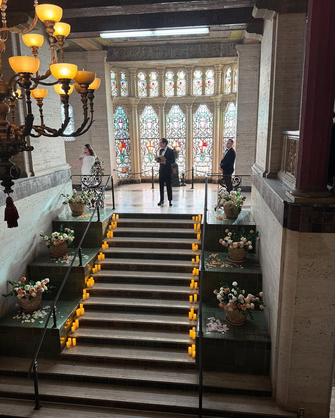 A wedding ceremony with a bride in a wedding dress and bouquet, a groom, and a officiant in a church decorated with flower arrangements and candles along the staircase, with stained glass windows in the background.