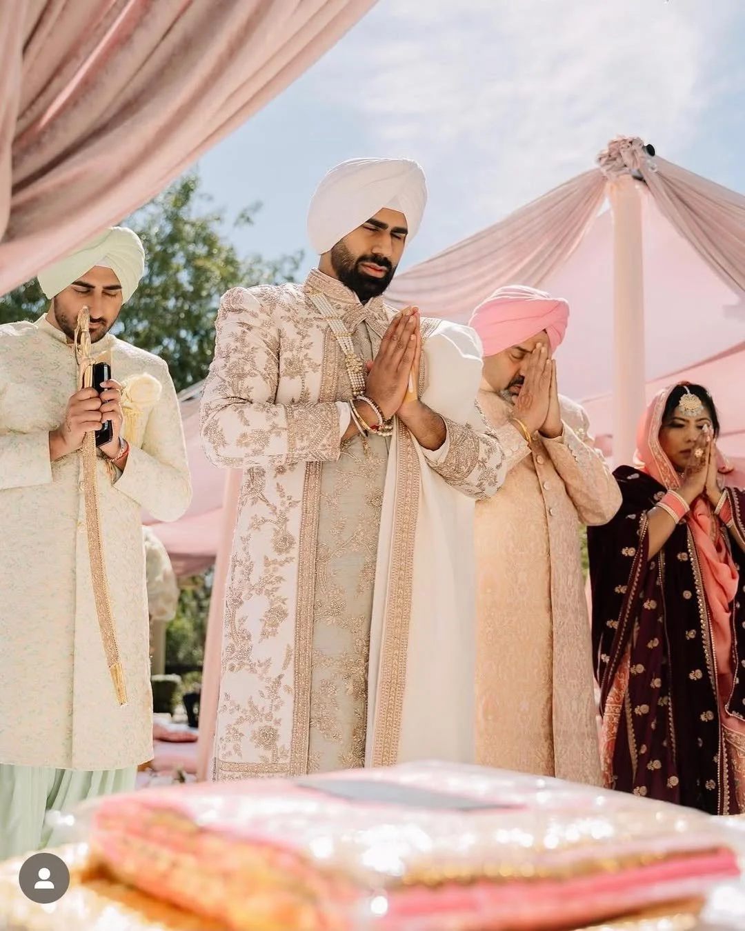 Group of people praying at an outdoor Indian wedding ceremony, with pink and white decorate tents in the background.