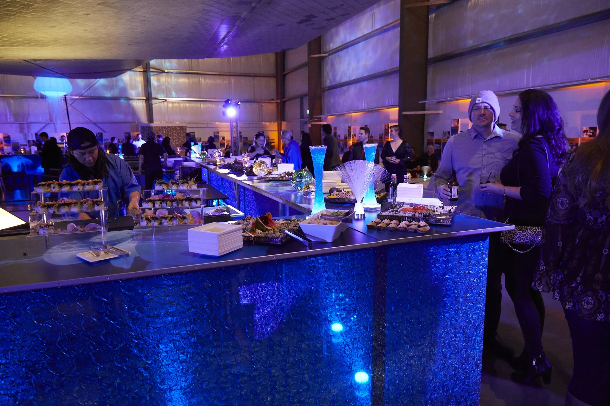 People socializing at a party with a decorated dessert table illuminated by colorful lights in a large indoor venue.