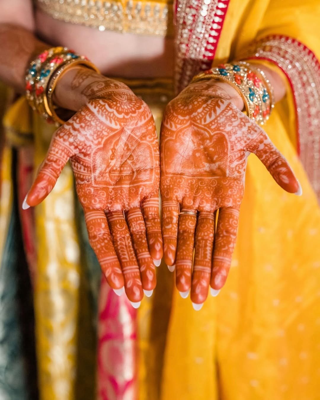 Close-up of hands with intricate henna designs, jewelry, and colorful traditional attire, likely for a celebration or wedding.