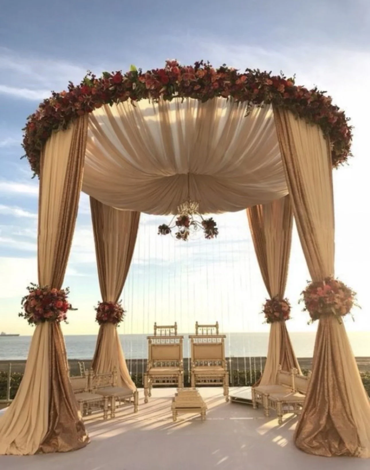 Elegant wedding altar with draped beige fabric, floral arrangements, and chairs, outdoors near the ocean at sunset.